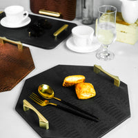 Isometric view of a black octagon serving tray with a spoon and two biscuits, showing the golden-colored handles from a different angle. Other trays are visible in the blurred background.