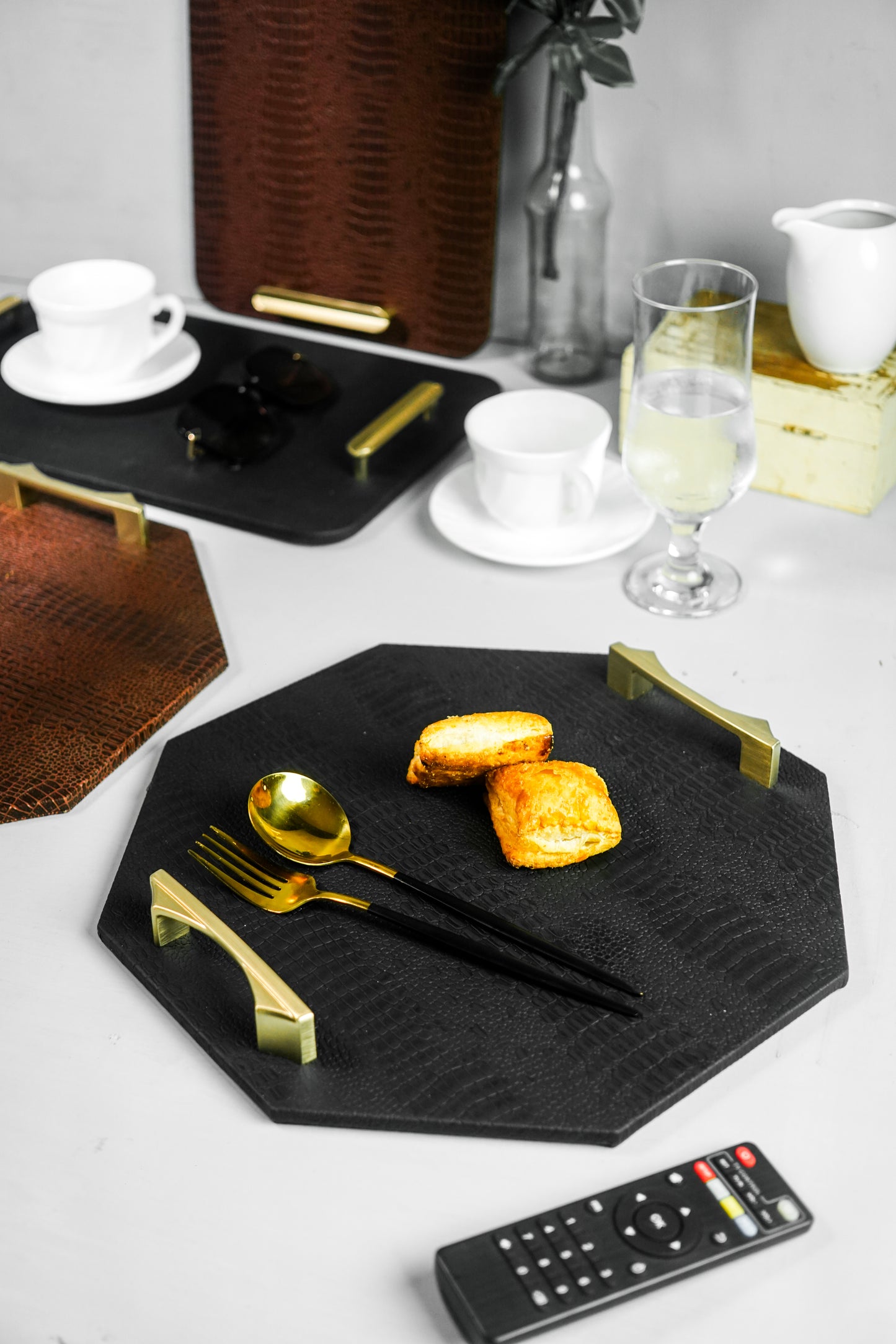 Isometric view of a black octagon serving tray with a spoon and two biscuits, showing the golden-colored handles from a different angle. Other trays are visible in the blurred background.