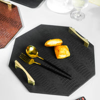 Image of a black octagon serving tray with a spoon and two biscuits on it, a remote on the side, and two blurred trays in the background with a glass and a tea cup on the sides.