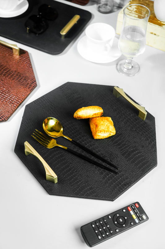 Image of a black octagon serving tray with a spoon and two biscuits on it, a remote on the side, and two blurred trays in the background with a glass and a tea cup on the sides.